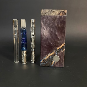 May include: A set of three glass and metal smoking pipes with a wooden box. The pipes are all silver with black lettering that says "Dynavap". The box is made of dark wood with a purple and blue swirl pattern.