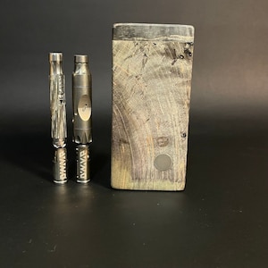 May include: A collection of vaping accessories, including two metallic vaporizers and a wooden storage case. The vaporizers have intricate designs and the case features a natural wood grain pattern with a circular button.