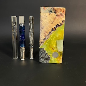 May include: An assortment of vaping accessories, including glass tubes, a blue glass piece, a metal piece, and a wooden box with a green and brown design. The items are arranged on a black surface.