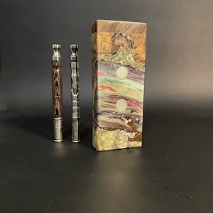 Può includere: Due pipe da fumo in metallo con dettagli in legno e una scatola di legno con un design marmoreo colorato. La scatola ha due cerchi argentati sul davanti.