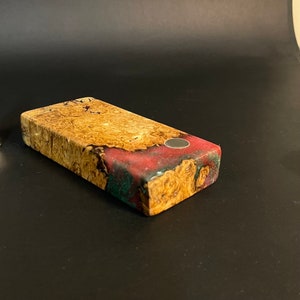 Op de afbeelding: Een rechthoekig houten blok met een rood, groen en bruin marmerpatroon. Het blok heeft een kleine, ronde, zilveren magneet die in het bovenoppervlak is ingebed.