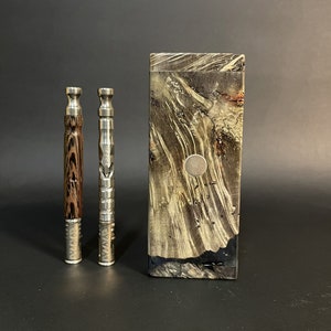 Può includere: Un set di tre articoli: due vaporizzatori cilindrici e una custodia rettangolare. Un vaporizzatore ha un motivo a venature del legno, l'altro è argentato. La custodia ha un aspetto marmorizzato con un bottone argentato.