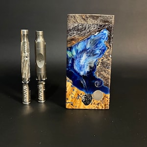 May include: Two silver metal vape pens with the text "AWVP" and a rectangular wooden box with a blue and white resin design. The box has a gold-colored wooden base and a black and silver top.