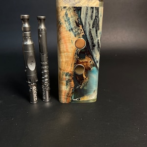 May include: Two black metal vape pens with the word "OMNI" engraved on them, standing in front of a wooden box with a blue, green, and brown marbled design.