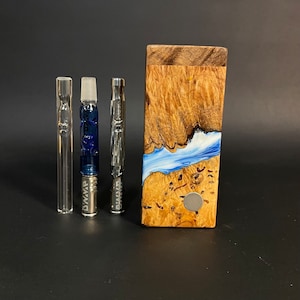 May include: An assortment of glass and metal smoking accessories, including a clear glass tube, a blue and clear glass piece, and a metal piece. A wooden box with a blue and white wave design is also present.