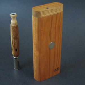 May include: A wooden dugout set with a matching one-hitter pipe. The dugout is rectangular, made of light brown wood, and has a small metal disc on the front. The one-hitter pipe is made of wood and metal.
