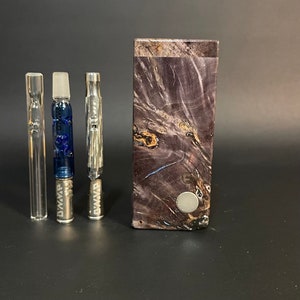 May include: Three silver metal vape cartridges with the word "DWAVP" engraved on them, next to a dark purple wooden box with a silver metal circle on the front.