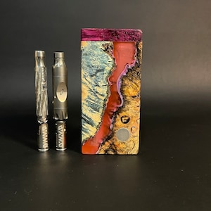 May include: A wooden box mod with a unique design, featuring a mix of red, purple, and natural wood tones. Two silver metal vaporizers are positioned to the left of the box mod. The box mod has a magnetic button and the letter "F" on the front.