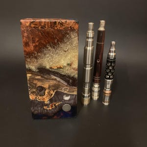 Könnte beinhalten: Eine rechteckige Holzkiste mit Harzfinish, in Braun-, Gold- und Blautönen. Drei Vape-Pens mit Metall- und Holzdetails befinden sich rechts. Die Stifte haben unterschiedliche Designs und Oberflächen.