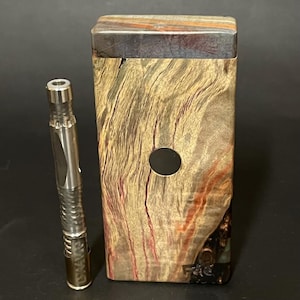 May include: A wooden box with a dark wood top and a circular hole in the front. The box has a wood grain pattern with red, brown, and black streaks. A silver metal tool with a textured surface is standing next to the box.