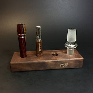 May include: A wooden stand with three slots for holding smoking accessories. The stand is made of dark wood and has the word "FUTO" engraved on the front. There are two wooden pipes and a glass bowl on the stand.