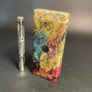 May include: A stainless steel vaporizer next to a rectangular wooden box. The box features a colorful, swirling pattern of green, yellow, red, and blue hues. A small, dark circle is centered on the box. The vaporizer is cylindrical with etched details.