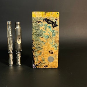 May include: Two silver metal tools with the word "Dynavap" engraved on them, next to a wooden box with a blue, green, and yellow marbled pattern.