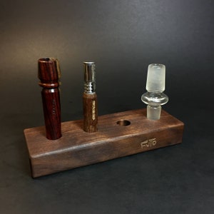 May include: A wooden stand with three slots for holding smoking accessories. The stand is made of dark wood and has the word "FUTO" engraved on the front. There are three accessories in the slots: a wooden pipe, a metal pipe, and a glass adapter.