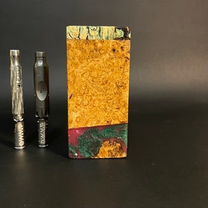 Op de afbeelding: Twee zwarte metalen vape-pennen met het woord "DWMA" erop gedrukt, staand voor een houten doos met een kleurrijk, abstract ontwerp. De doos heeft een groen, blauw en paars patroon.