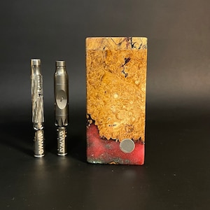 Op de afbeelding: Twee zilveren metalen rookpijpen met het woord "Dynavap" erop gegraveerd, naast een houten doos met een rood-bruin marmerpatroon.