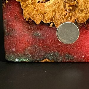 Op de afbeelding: Een close-up van een rood, groen en bruin kunsthars kunstwerk met een rond, grijs metalen accent. Het hars heeft een glinsterende, glimmende textuur.