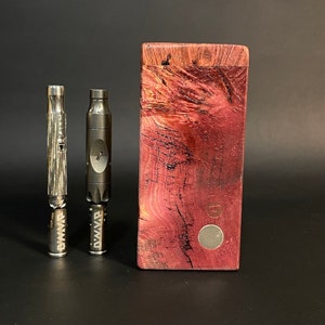 May include: Two silver metal vaporizers with the word "DWMAV" engraved on them, standing on a black surface in front of a wooden box with a purple and brown wood grain pattern.