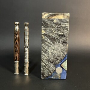 Può includere: Due vaporizzatori con dettagli in metallo e legno sono posizionati accanto a una scatola rettangolare con un motivo unico di venature del legno. I vaporizzatori hanno un design elegante e cilindrico. La scatola presenta un mix di tonalità grigie, nere e blu.