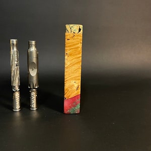 Op de afbeelding: Twee zilveren metalen rookpijpen met het woord "DWVR" erop gegraveerd, en een houten blok met een rode en groene harsinleg.