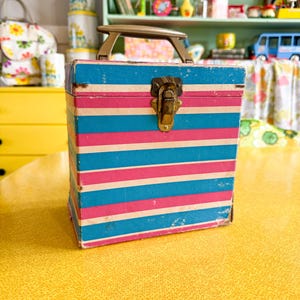 Puede incluir: Una caja vintage con asa, con rayas horizontales en azul, rosa y blanco. La caja tiene un cierre metálico y un asa marrón. La caja está sobre una superficie amarilla.