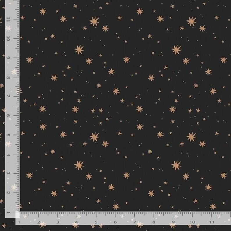 May include: Black fabric with a repeating pattern of gold-colored stars and tiny white dots. The stars vary in size and shape, creating a celestial design. A ruler is visible along the left and bottom edges, marked in inches.