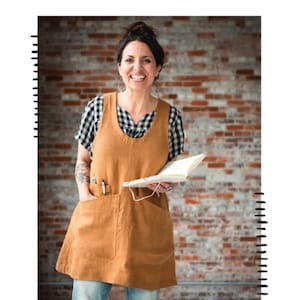 May include: A woman wearing a brown studio tunic over a black and white checkered shirt, holding a notebook. The tunic has pockets and the text "STUDIO TUNIC" is at the bottom. The top of the image has the text "SEW LIBERATED" and a scissors logo.
