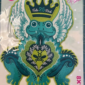 May include: Embroidered patch featuring a teal frog with a crown and wings. The patch includes the text "Tula 25 Pink" and measures 8 x 10 inches. The design is a limited edition.