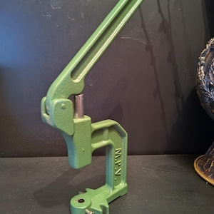 May include: A green metal press with a long handle and a base with the letters "M.P.V." stamped on the side. The press is designed for applying pressure, likely for crafting or industrial use. The press is painted in a solid green color.