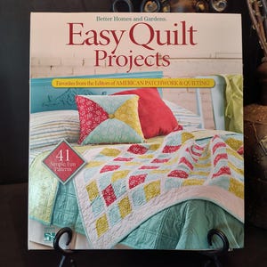 May include: A book titled "Easy Quilt Projects" by Better Homes and Gardens, featuring a colorful quilt and pillows. The cover displays the text "41 Simple Fun Patterns" and "Favorites from the Editors of AMERICAN PATCHWORK & QUILTING". The book is displayed on a black stand.
