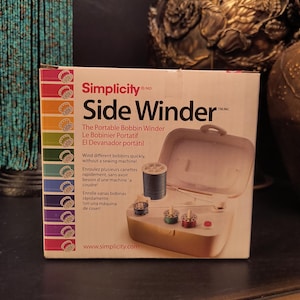 May include: A Simplicity Side Winder box, a portable bobbin winder. The box is white with a colorful stripe on the left side. The winder is beige with a clear lid, holding spools of thread.