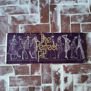 May include: A vintage book cover with a purple background and the text "The Perfect Fit" in gold lettering. The cover features illustrations of people wearing different clothing styles. The text below the title reads "Create over 1200 perfect fitting, fashionable styles in your own home, with the exclusive "Over Lay" and "Trans-Dart" method."
