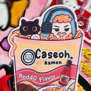 Könnte beinhalten: Ein farbenfroher Aufkleber mit einer Cartoon-Ramen-Schale mit dem Text "Caseoh. Ramen Red40 Flavor". Der Aufkleber enthält Illustrationen einer Katze, einer Person mit Kopfhörern und einer Flasche scharfer Soße. Der Hintergrund ist rosa.