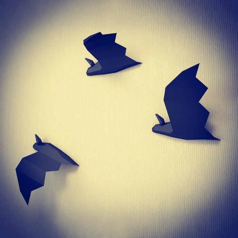 May include: Three black paper bats are mounted on a light beige wall. The bats are in different positions, with one flying upwards, one flying downwards, and one flying to the right.