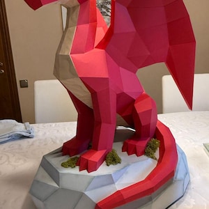 May include: A red paper dragon sculpture with geometric shapes, perched on a grey, polygonal base with green moss accents. The dragon has a long tail and wings, and a light green eye. A unique, handcrafted fantasy art piece.