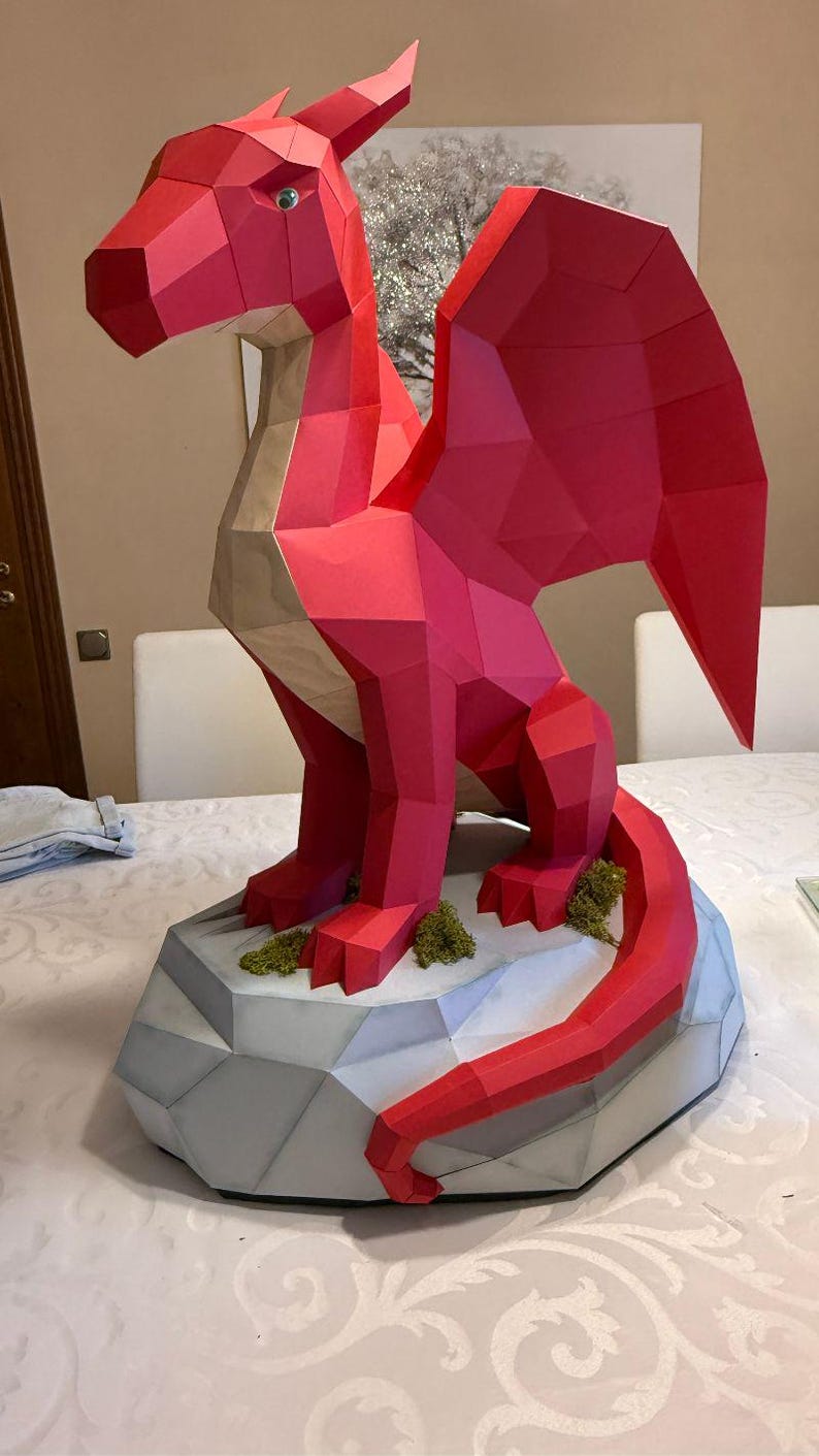 May include: A red paper dragon sculpture with geometric shapes, perched on a grey, polygonal base with green moss accents. The dragon has a long tail and wings, and a light green eye. A unique, handcrafted fantasy art piece.