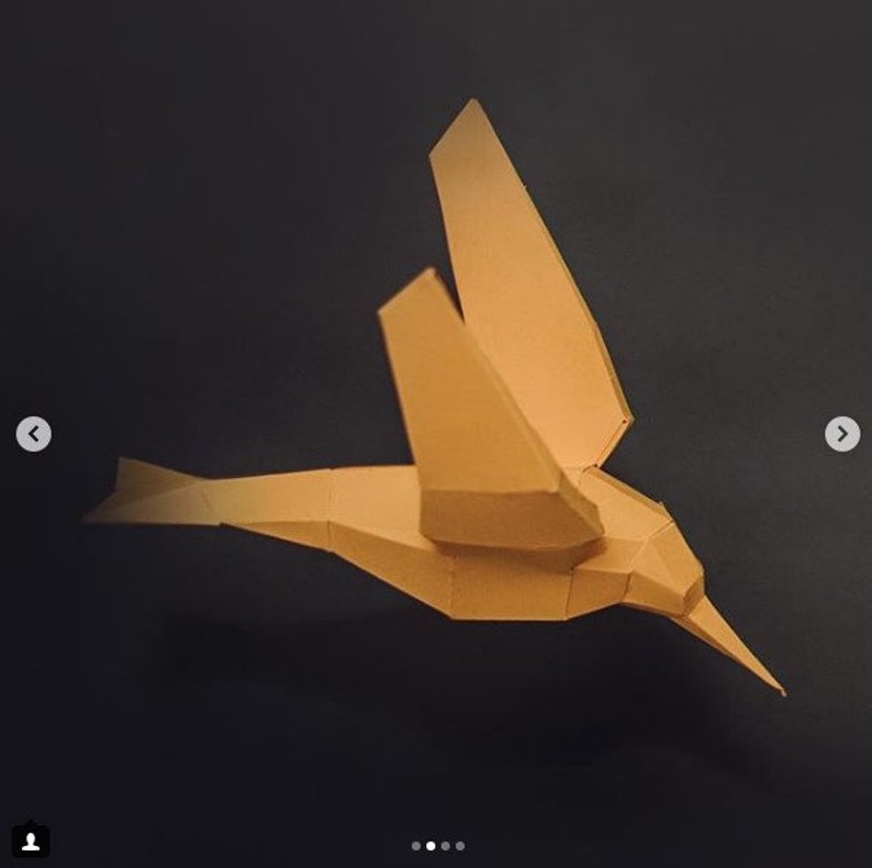 May include: A tan paper origami hummingbird sculpture with a long beak and wings folded back. The sculpture is on a black background.