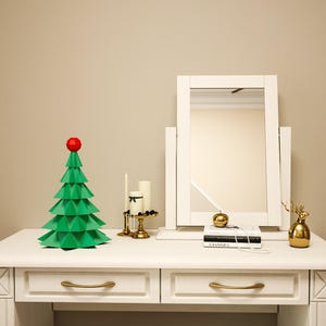 May include: A white vanity table with two drawers and gold handles. A green origami Christmas tree with a red star on top sits on the table. A white framed mirror with a gold accent sits behind the tree. A gold decorative deer figurine and a book with the word "CHANEL" on the cover are also on the table.