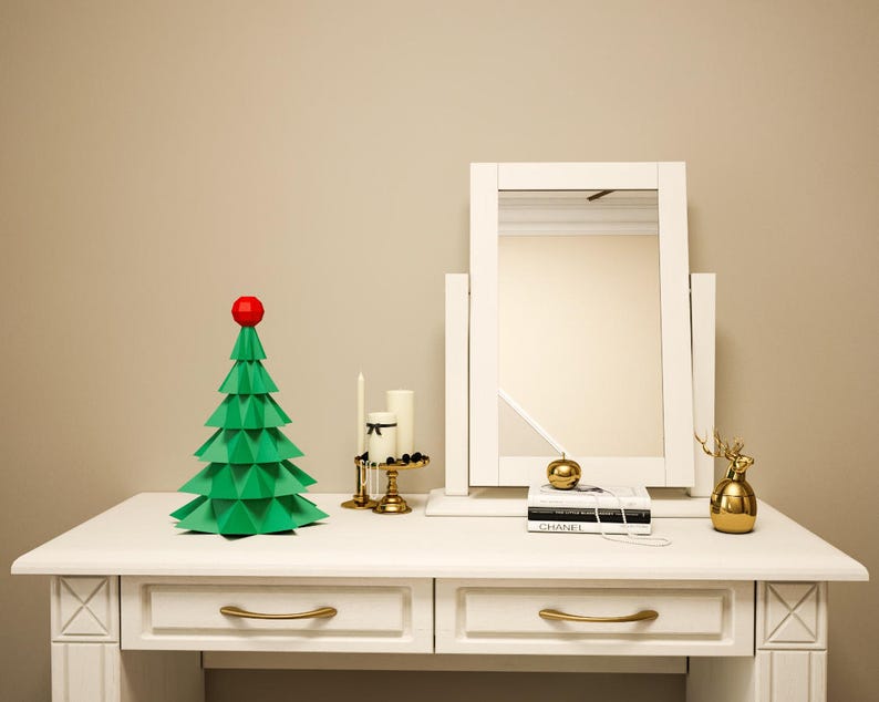 May include: A white vanity table with two drawers and gold handles. A green origami Christmas tree with a red star on top sits on the table. A white framed mirror with a gold accent sits behind the tree. A gold decorative deer figurine and a book with the word "CHANEL" on the cover are also on the table.