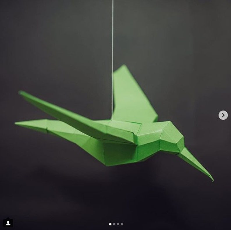 May include: A green paper hummingbird mobile hanging from a white string against a gray background.