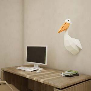 May include: A white origami pelican wall sculpture with an orange beak, mounted on a light beige wall above a wooden desk with a computer monitor, keyboard, and mouse.
