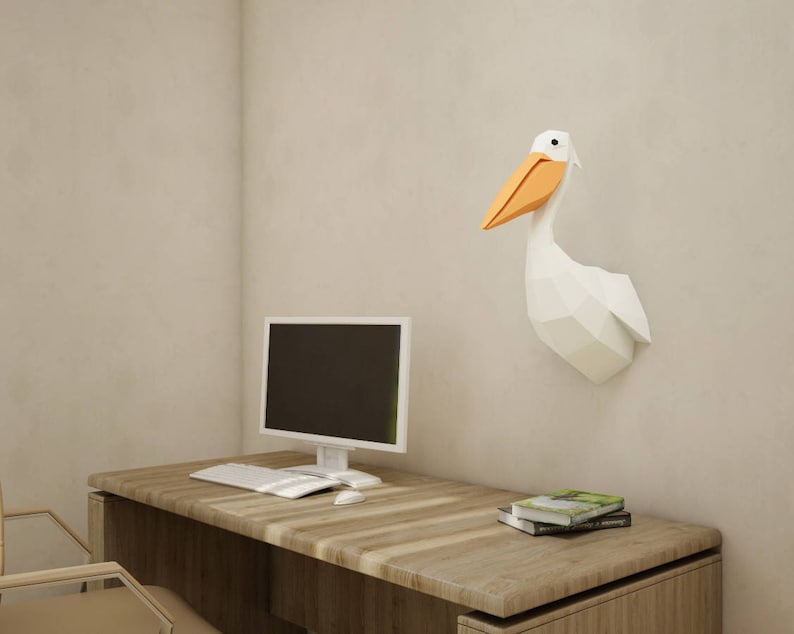 May include: A white origami pelican wall sculpture with an orange beak, mounted on a light beige wall above a wooden desk with a computer monitor, keyboard, and mouse.