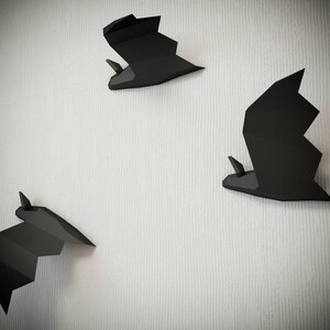 May include: Three black paper bats are mounted on a white textured wall. The bats are in different sizes and positions.