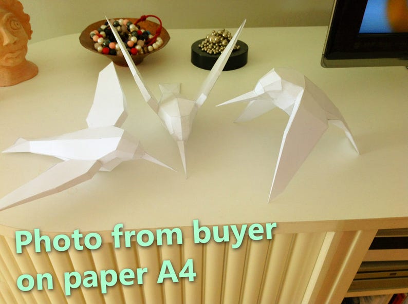 May include: Three white paper origami birds, a hummingbird, a crane, and a dove, are displayed on a white surface. The birds are made from A4 paper. "Photo from buyer on paper A4" is written on the surface below the birds.