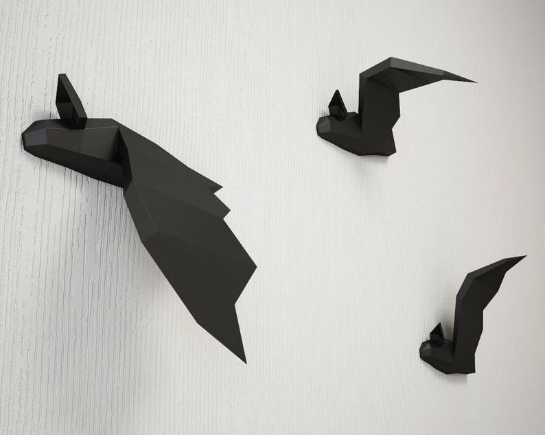 May include: Three black paper bats are mounted on a white wall. The bats are made of folded paper and have a 3D effect.