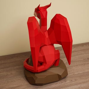 May include: A red, geometric paper dragon sculpture with outstretched wings and a detailed form. The dragon is perched on a brown, polygonal base, creating a striking contrast against the wooden surface.
