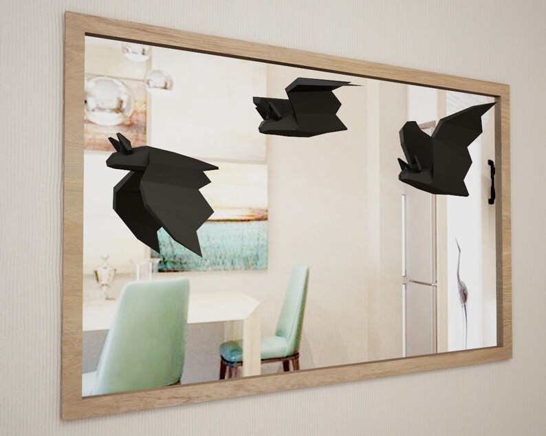 May include: Three black origami bats are hanging on a wall mirror. The bats are in flight position with their wings spread out. The mirror is framed in wood and reflects a room with furniture.