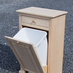 May include: A light wood trash can with a white plastic liner. The trash can has a pull-out drawer and a hinged door. The door is open and the liner is partially visible. The trash can is sitting on a gravel surface.