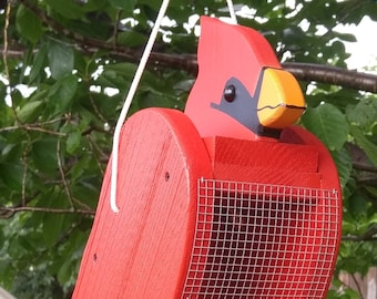 May include: Red wooden cardinal bird feeder with a wire mesh seed tray and a hanging loop. The feeder is painted red with black eyes and a yellow beak.