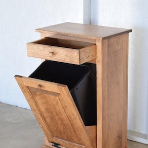 May include: A wooden trash can cabinet with a single drawer and a pull-out door. The cabinet is made of light brown wood and has black metal hinges.